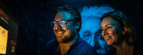 Deux adultes souriants, un homme barbu aux lunettes et une femme, observant un écran de jeu avec le mot "EINSTEIN" et un score de "18", dans un environnement sombre et immersif avec un grand portrait bleu d'Einstein en arrière-plan chez Prison Island 77. Cette image illustre des activités à faire intellectuelles et collaboratives, idéales pour une sortie de loisirs, un team building, une journée de cohésion, un anniversaire, un EVG ou un EVJF immersif entre amis ou en famille en Seine-et-Marne, à Émerainville (Marne-la-Vallée), proche de Pontault-Combault, dans l'esprit des défis de Fort Boyard.