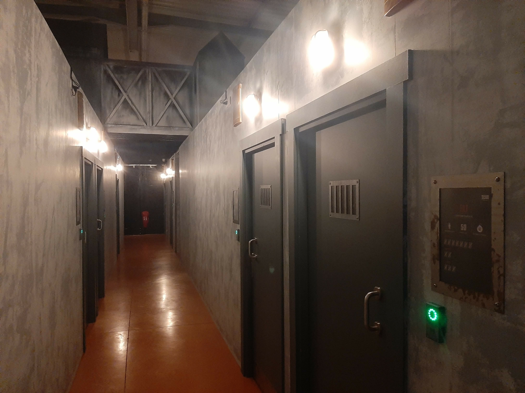 Un long couloir sombre bordé de portes de cellules de prison métalliques et de lumières murales, évoquant l'ambiance immersive des activités à faire chez Prison Island 77 à Émerainville. Cette image illustre parfaitement une sortie de loisirs originale et captivante entre amis ou en famille en Seine-et-Marne, dans l'esprit des défis de Fort Boyard.