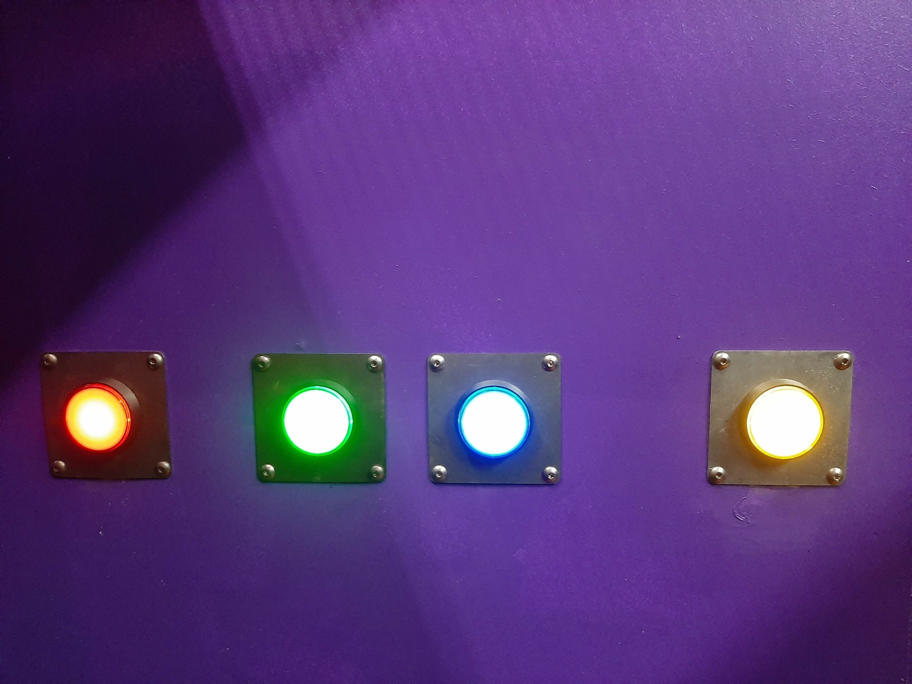 Une série de boutons lumineux de différentes couleurs (rouge, vert, bleu, jaune) sur un mur violet, illustrant les défis interactifs et les activités à faire pour une sortie de loisirs stimulante chez Prison Island 77 en Seine-et-Marne, parfait entre amis ou en famille, dans l'esprit de Fort Boyard.