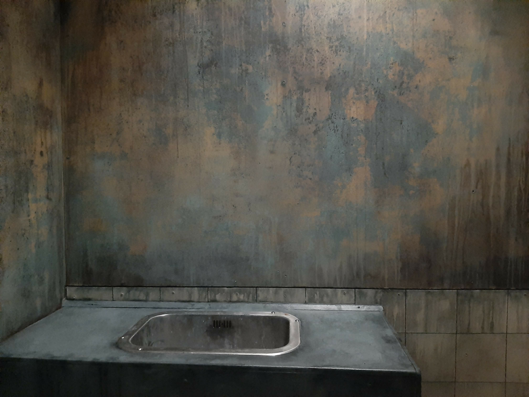 Un coin de pièce à l'aspect vieilli et industriel, avec un mur texturé aux tons rouille et bleu-gris, et un évier métallique encastré sur un comptoir sombre, évoquant l'ambiance immersive de Prison Island 77 et les activités à faire lors d'une sortie de loisirs unique entre amis ou en famille en Seine-et-Marne, dans l'esprit de Fort Boyard.