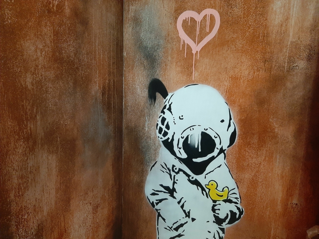Une œuvre murale de style graffiti représentant un personnage avec un casque de scaphandre tenant un canard jaune, avec un cœur rose dégoulinant au-dessus, illustrant l'ambiance artistique et les activités à faire lors d'une sortie de loisirs unique chez Prison Island 77 en Seine-et-Marne, rappelant l'esprit de Fort Boyard, idéal entre amis ou en famille.