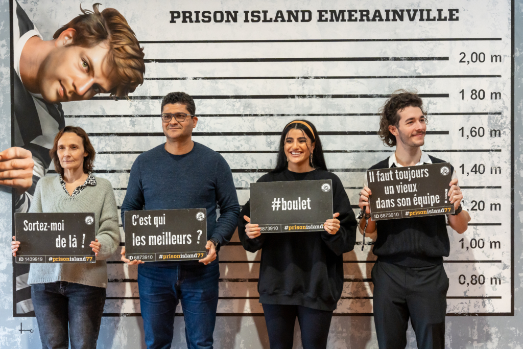 Quatre personnes, dont une femme plus âgée, un homme d'âge moyen, et deux jeunes adultes, posent devant une toise de prisonnier géante avec humour, tenant des pancartes.