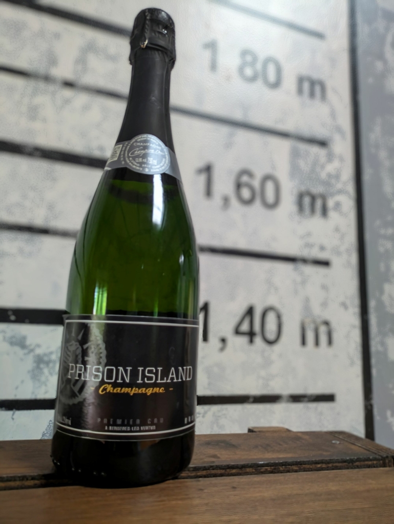 Bouteille de champagne personnalisée "Prison Island Champagne" devant un fond mesurant des hauteurs, symbolisant la célébration après une évasion réussie.